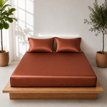 Bed with rust-colored bedding and pillows in a room with plants and light streaming through a window.