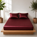 A bedroom with a wood platform bed, beautiful maroon satin sheets and pillows. Potted plants add to the peace and coziness of a room.