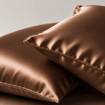 Two satin pillowcases in a warm metallic bronze color, shown close-up against a neutral light gray background.