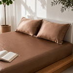 Close-up of a light brown satin bedsheet with two matching pillow covers on a wooden bed with an open book