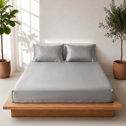 A neatly made bed with a light grey fitted sheet and two matching pillowcases on a low wooden platform bed, styled in a bright room with potted plants and soft window shadows.