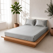 Side angle of a silver grey satin bedsheet with matching pillow covers on a wooden platform bed beside indoor plants