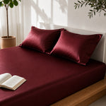 Burgundy bed sheets with pillows and an open book on a bed, with plants in the background.