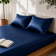 A neatly made bed with a dark navy blue bedsheet and matching pillowcases, styled in a bright bedroom with a wooden headboard, a potted plant, and an open book on the bed.