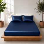 A navy blue satin sheet set with two matching pillowcases on a low wooden platform bed in a bright, modern bedroom with potted plants.