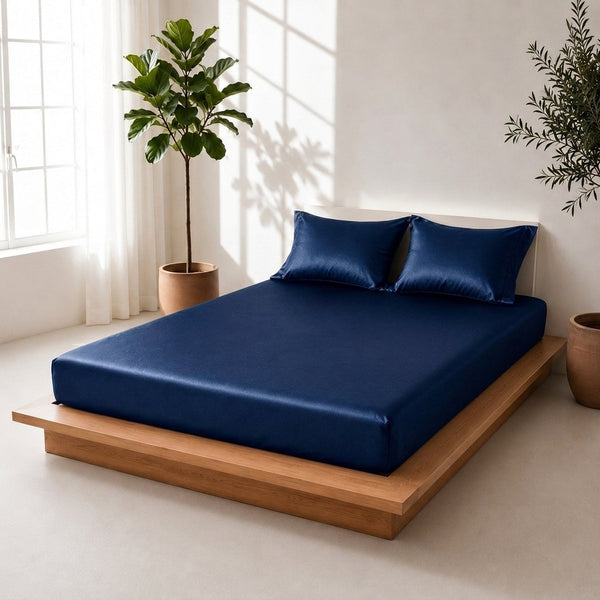 A navy blue bed sheet set with two matching pillowcases on a wooden platform bed in a bright, modern bedroom with potted plants.