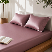 Rose satin sheets on a bed with pillows and an open book, in a bright room with plants.
