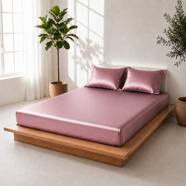 Pink satin bedding set on a wooden platform bed with plants in the background.