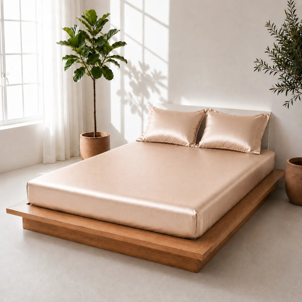 A bed dressed in a shiny sand colored satin sheet and two matching pillows, set on a low wooden platform bed in a bright, modern room with potted plants and natural window light.