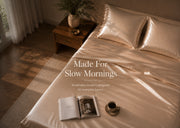 A bed dressed in shiny champagne-colored satin bedding with two matching pillowcases, styled in a warm bedroom scene with a wooden nightstand, curtains, and overlaid text reading 'Made For Slow Mornings'.